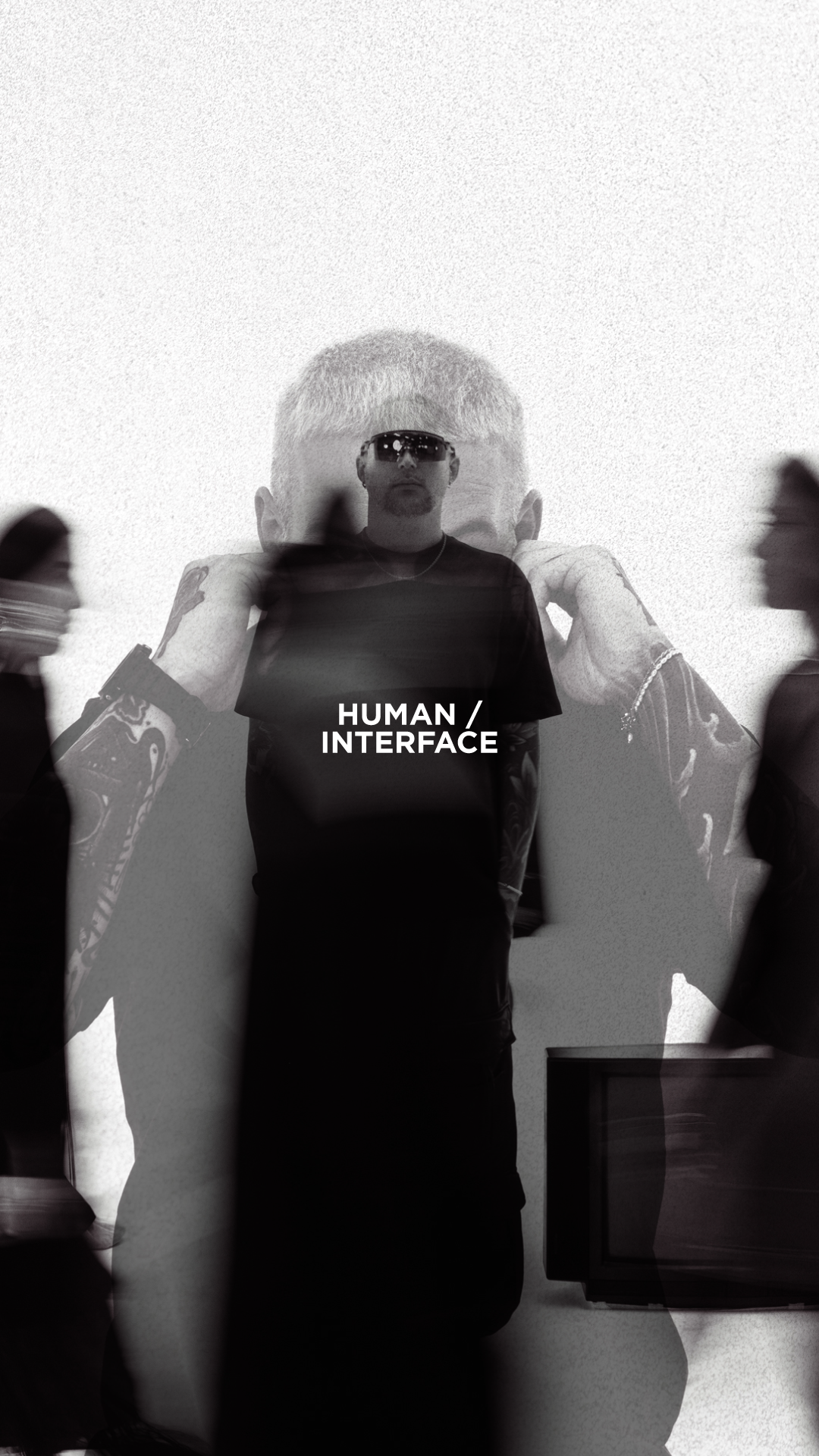 Persona con capelli bianchi, occhiali da sole e t-shirt nera con scritta 'HUMAN/INTERFACE' circondato da altre persone